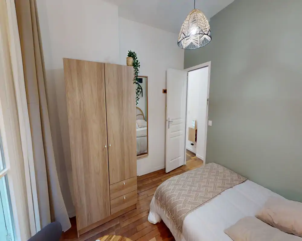Chambre lumineuse et chaleureuse avec lit double partiellement visible au premier plan, armoire en bois, miroir en pied et parquet chaleureux — parfaite pour une annonce de location.