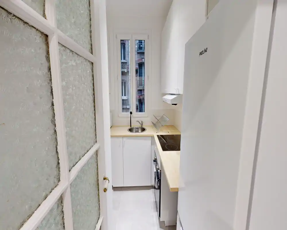 Cucina compatta e moderna con mobili bianchi, piano in legno, piano cottura elettrico integrato e lavabo tondo sotto una finestra alta che illumina naturalmente.