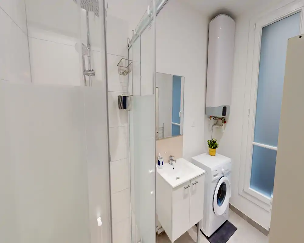 Bagno moderno compatto con box doccia, scaldabagno a parete, piccolo lavabo con mobiletto e lavatrice; finiture bianche e finestra satinata che donano un aspetto pulito e funzionale.