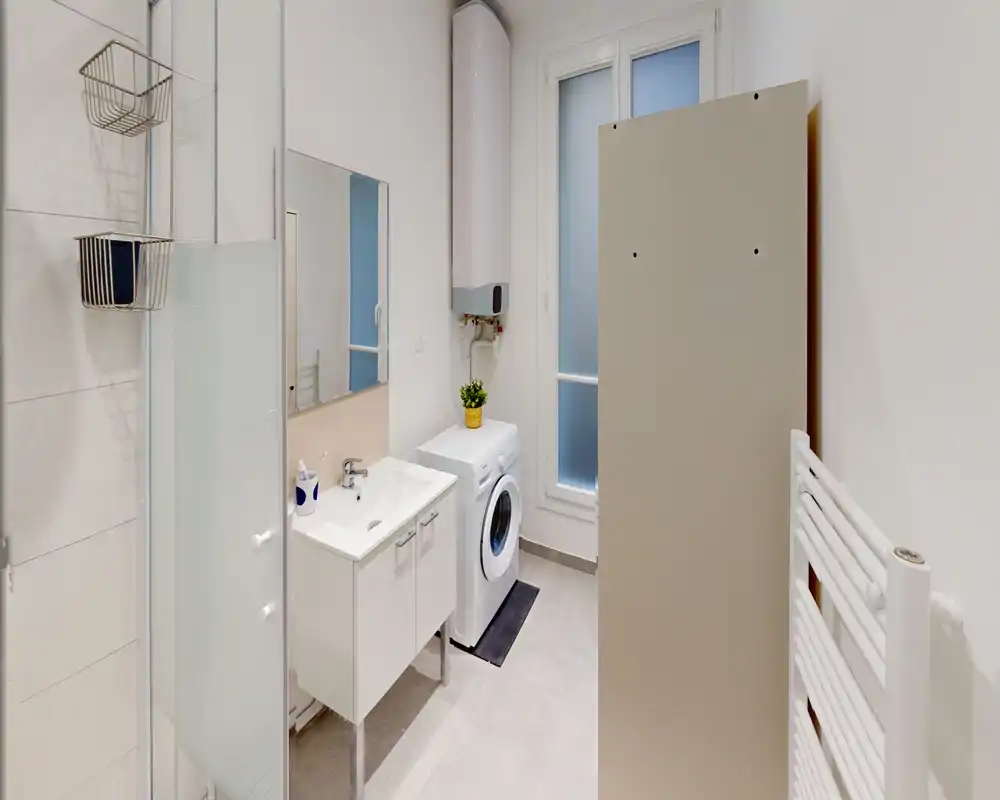 Bagno moderno e luminoso con mobile lavabo compatto, lavatrice a carica frontale, scaldabagno verticale, parete doccia in vetro e termoarredo — finiture neutre e ottima illuminazione.
