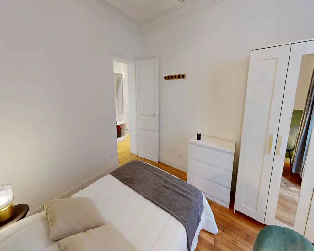 Chambre lumineuse et soignée avec lit double, textiles doux et parquet chaleureux. Armoire et commode blanches, tons neutres apportant une atmosphère reposante.