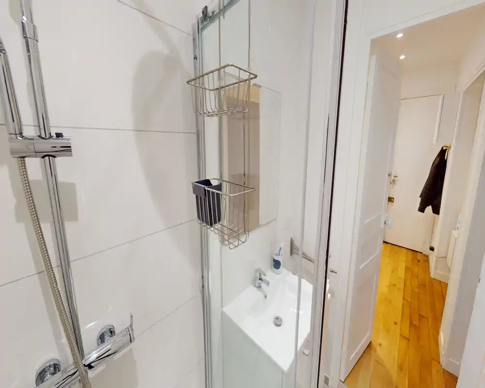 Salle de bains moderne et compacte avec paroi de douche en verre, robinetterie chromée et petit lavabo ; carrelage blanc lumineux et couloir en bois visible par la porte ouverte.