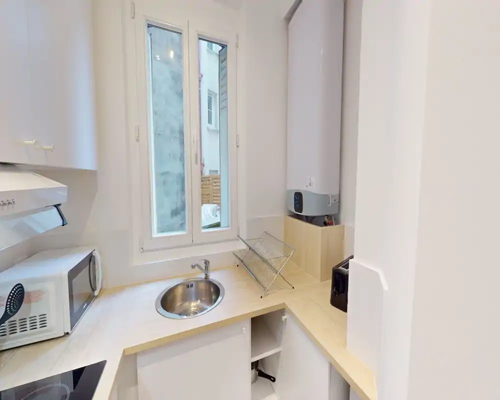 Kitchenette moderne et compacte avec plans de travail en bois clair, évier rond en inox sous la fenêtre, micro-ondes et petits appareils — lumineuse et bien organisée pour la vie urbaine.