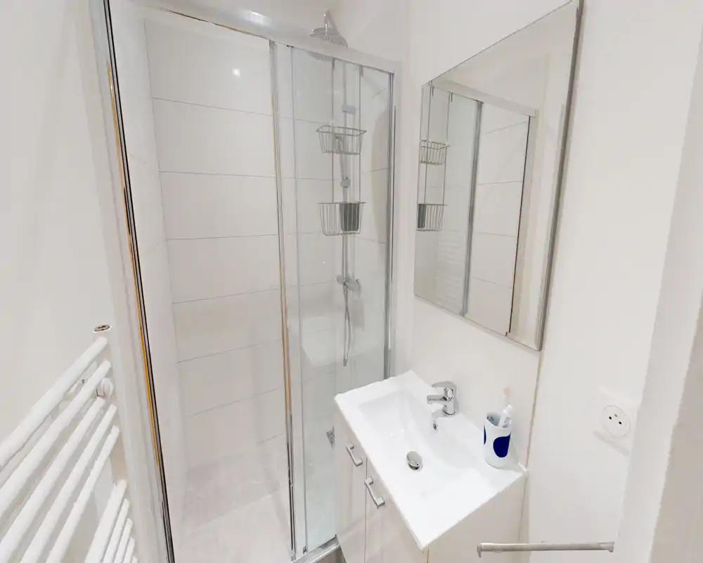 Salle de bains moderne et compacte avec douche vitrée, meuble lavabo blanc et armoire miroir — ambiance lumineuse et propre, idéale pour les locations courte durée.