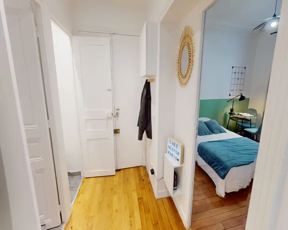 Entrée d'appartement lumineuse avec parquet et porte d'entrée blanche ; une ouverture latérale laisse voir la chambre. Couloir propre et fonctionnel, idéal pour accueillir des invités.