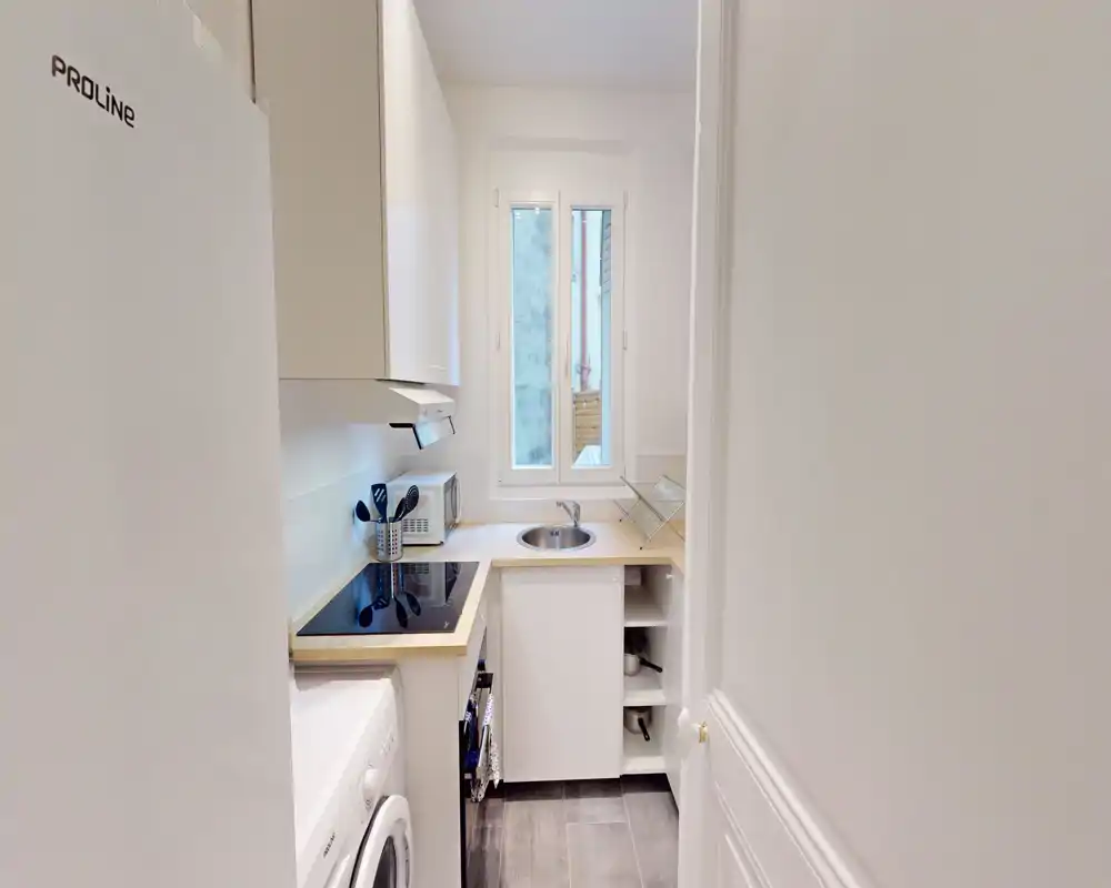 Cuisine compacte et moderne avec plaque à induction, évier rond sous la fenêtre et plans de travail en bois clair — idéale pour un petit appartement.