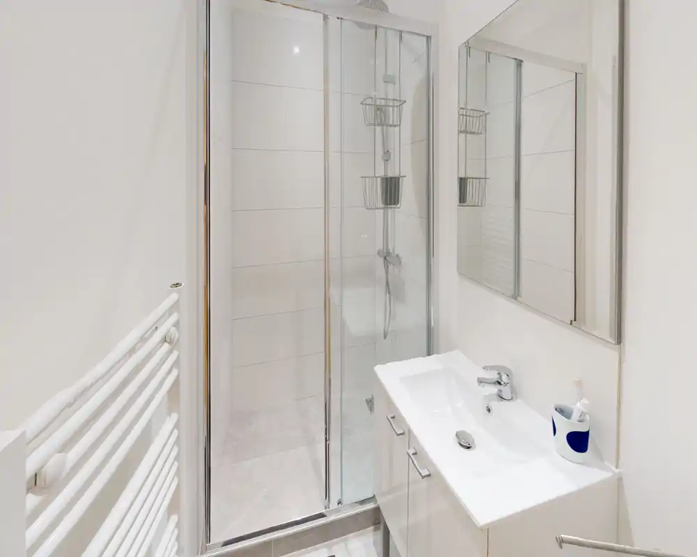 Salle de bains moderne et lumineuse avec douche à paroi vitrée, miroir mural et meuble-vasque compact — carrelage blanc et robinetterie chromée pour une impression de fraîcheur.