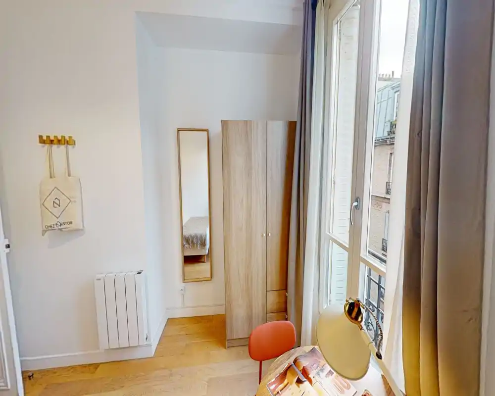 Rincón de dormitorio luminoso y ordenado con armario, espejo de cuerpo entero y un vistazo de la cama; ventana grande con mucha luz natural y suelos de madera cálidos.