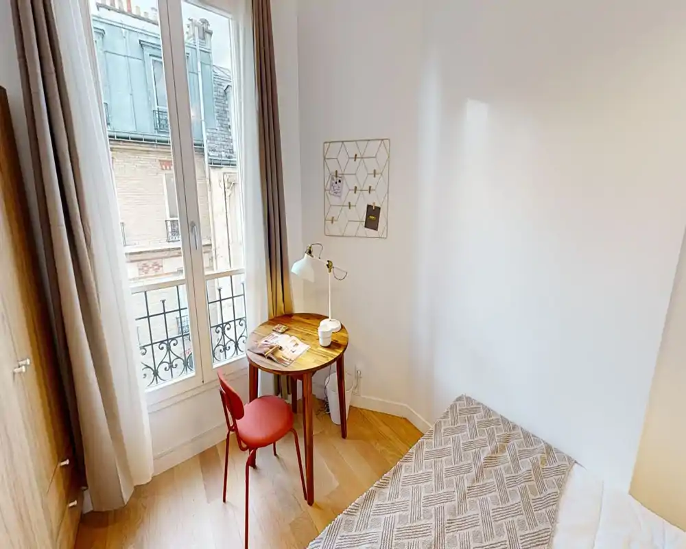 Pequeña habitación luminosa con cama individual, suelo de parquet y ventana que aporta mucha luz natural; una mesa redonda de madera con una silla roja crea un rincón de trabajo acogedor.
