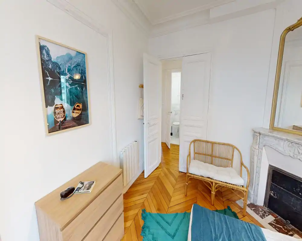Rincón de dormitorio luminoso y con estilo con suelo de espiga de madera, un banco de ratán, cómoda y vista parcial de la cama; molduras blancas y chimenea de mármol añaden carácter.