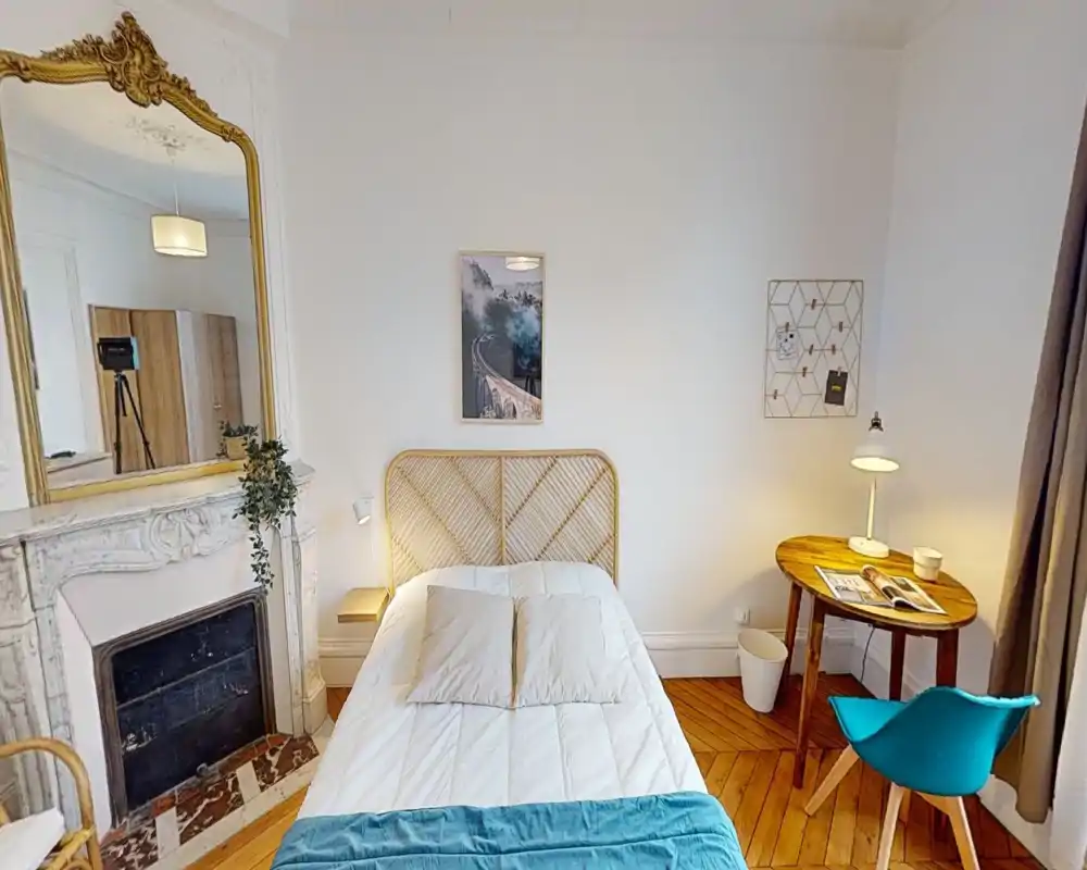 Dormitorio luminoso y con estilo con cama centrada y cabecero de rattan, espejo decorativo, suelo de parquet y mesita con lámpara.