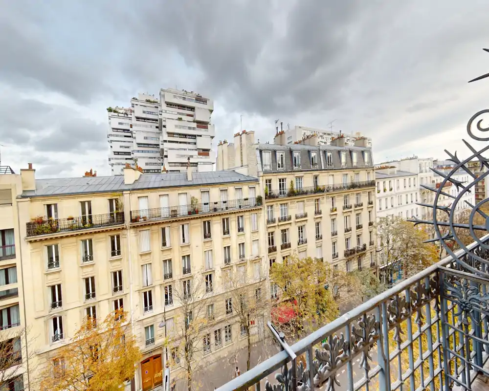 Vistas desde el balcón hacia edificios parisinos clásicos y una calle arbolada, con una barandilla de hierro forjado ornamentada y un cielo nublado.