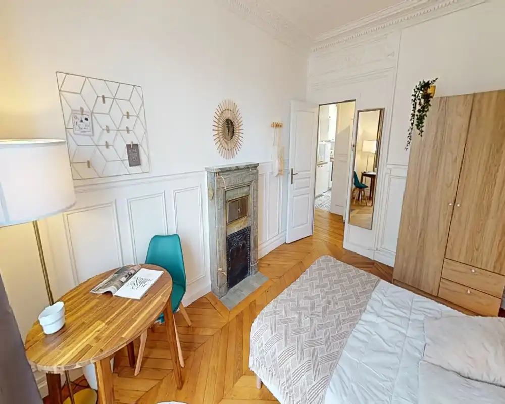 Dormitorio luminoso y con estilo, con cama doble, suelo de parquet, chimenea decorativa y una pequeña mesa — acogedor y bien iluminado para alquileres de corta estancia.