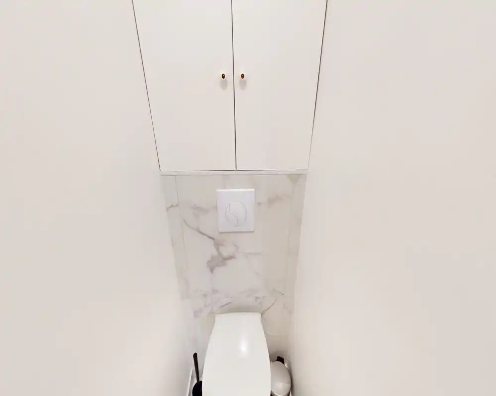 Aseo compacto y moderno con pulsador de cisterna empotrado, panel efecto mármol, acabados blancos y armario alto sobre el inodoro.