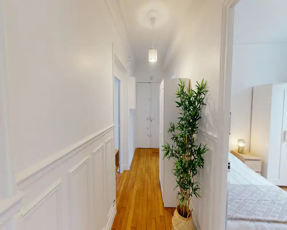 Entrada luminosa y estrecha con suelos de madera, zócalo blanco y una planta decorativa; se ve un dormitorio por la puerta de la derecha.