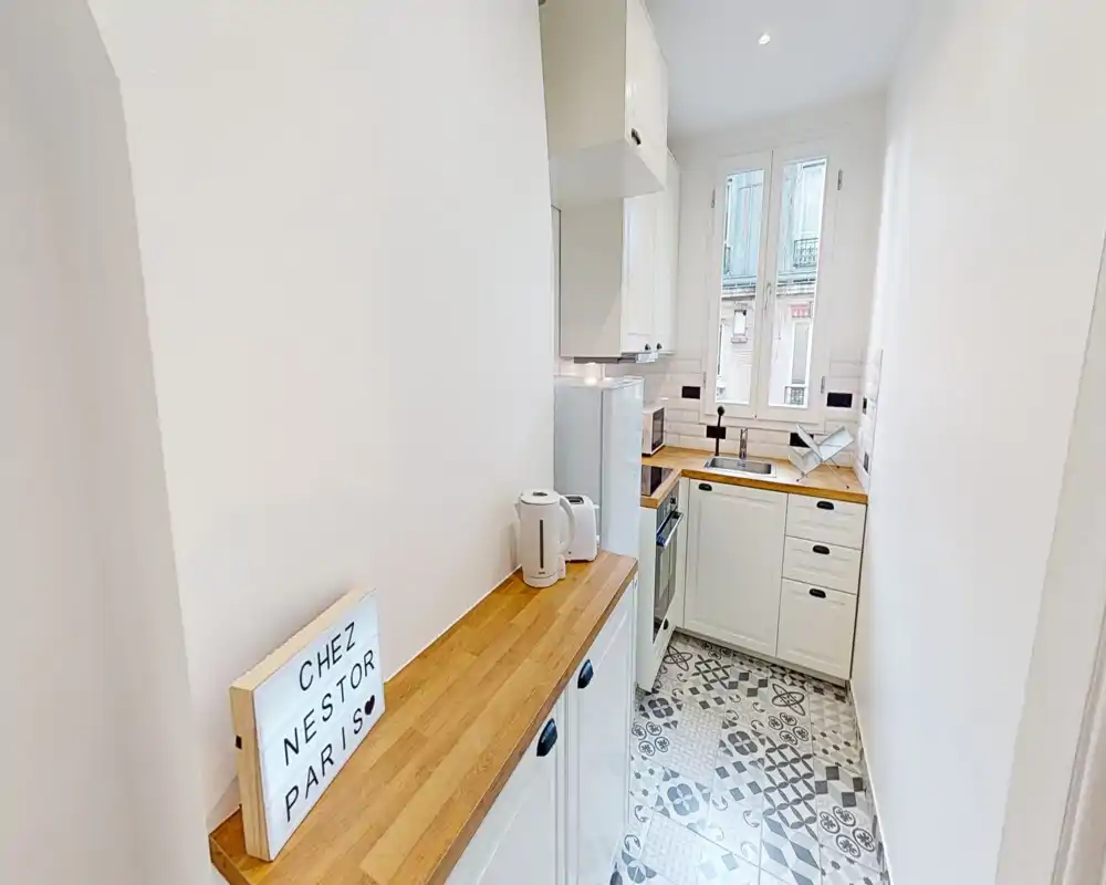 Cocina compacta y luminosa con encimeras de madera, suelo de baldosas estampadas y ventana que aporta luz natural — distribución ordenada ideal para un apartamento pequeño.
