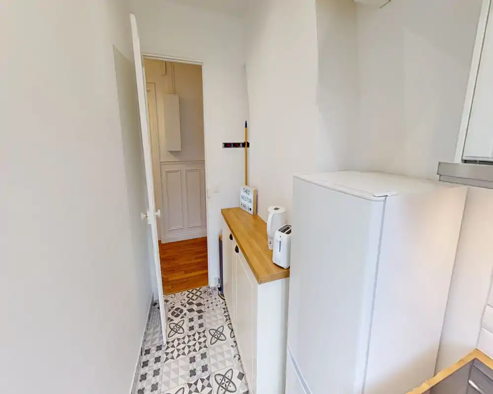 Cocina estrecha en forma de pasillo con suelo de baldosas decorativas, encimera de madera y pequeños electrodomésticos (hervidor, tostadora) junto a un frigorífico alto — espacio luminoso y funcional para pisos pequeños.