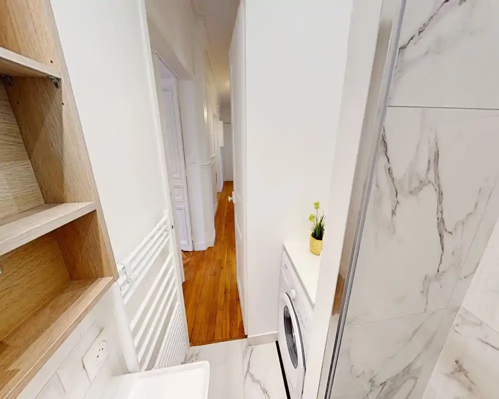 Rincón de baño pequeño con azulejos efecto mármol, puerta de ducha a la derecha y lavadora integrada bajo la encimera; distribución compacta que da a un pasillo.