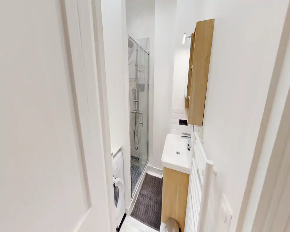 Baño moderno y compacto con mampara de ducha de vidrio, mueble de lavabo mural y toallero radiador. Paredes blancas y distribución funcional, ideal para un piso pequeño.