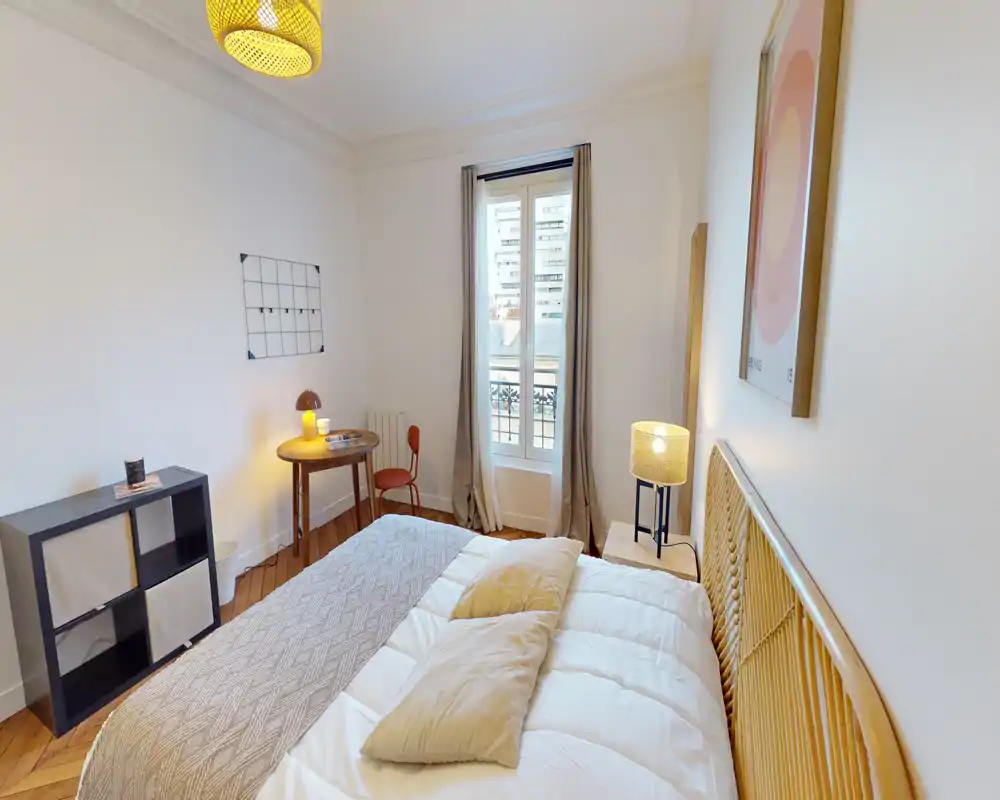Camera da letto luminosa e accogliente con testiera in rattan, biancheria comoda e lampade da comodino calde. Un piccolo scrittoio e la finestra con tende offrono luce naturale e funzionalità.