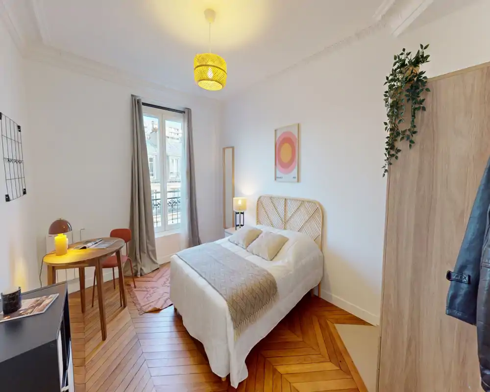 Camera da letto luminosa e accogliente con un comodo letto matrimoniale, biancheria morbida, pavimento in parquet e una finestra che illumina naturalmente. Dettagli caldi come lampada, tappeto e quadro rendono l'ambiente invitante.