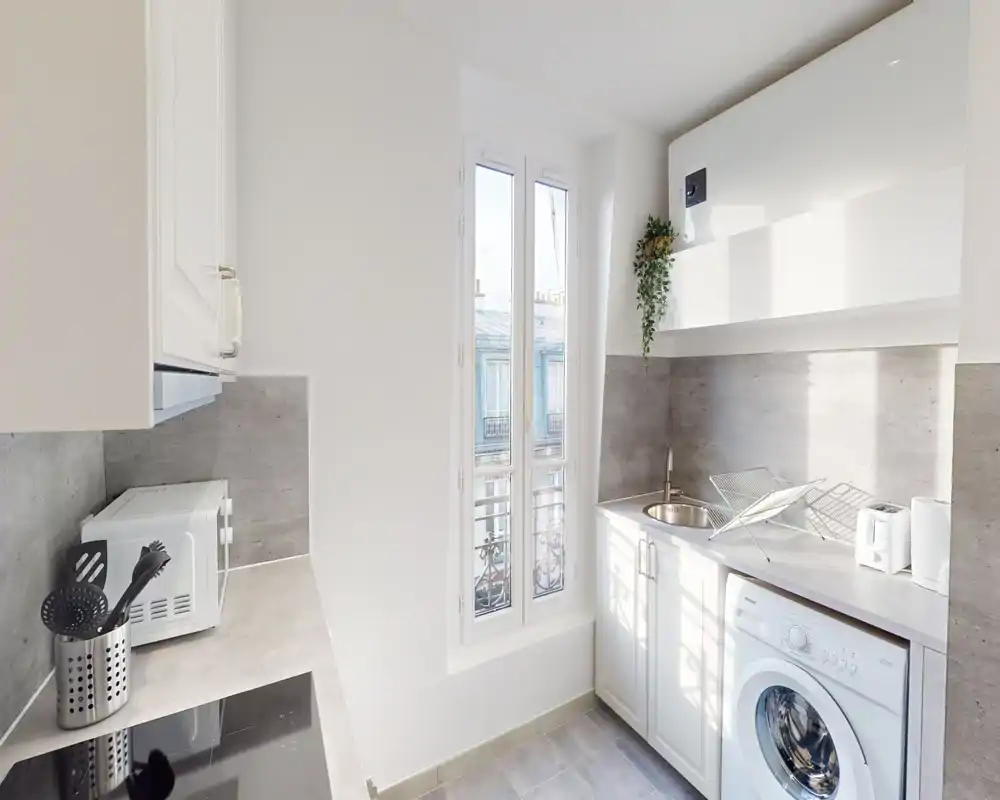 Cucina compatta e luminosa con pensili bianchi, lavello, piccoli elettrodomestici sul piano e una lavatrice accanto a una finestra che illumina l'ambiente.