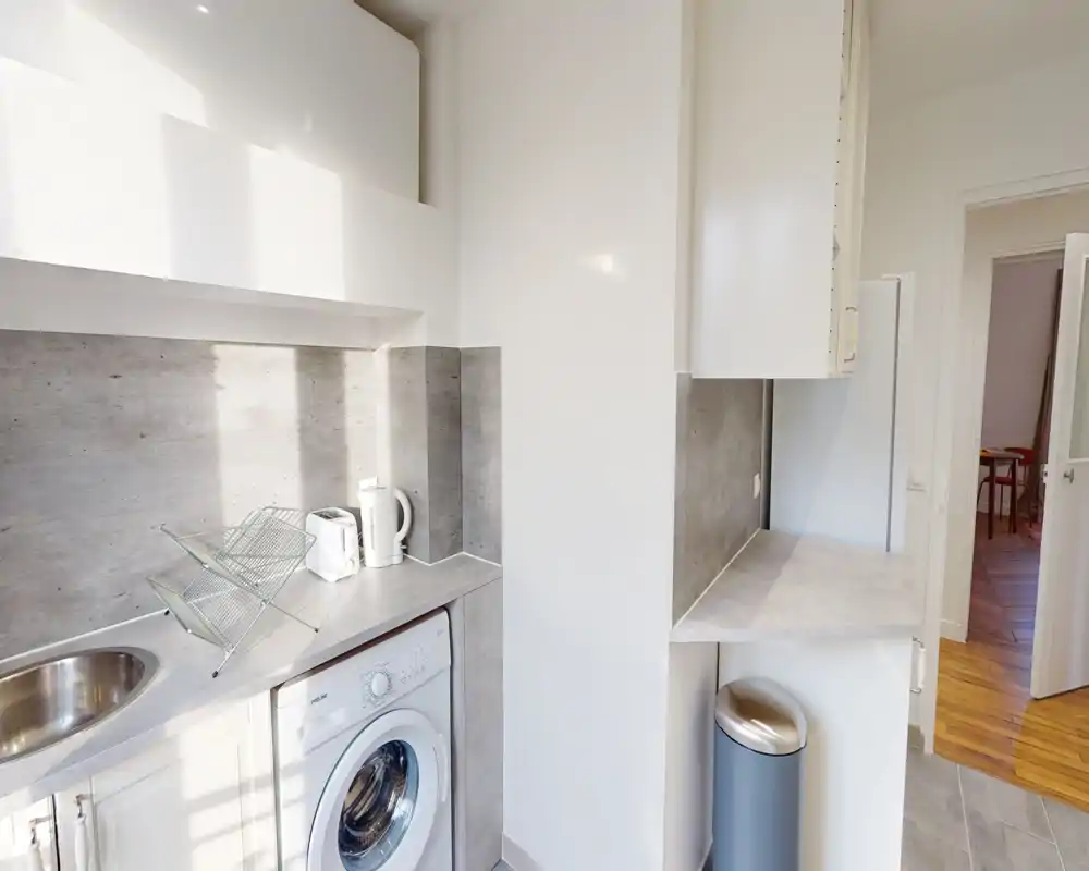 Angolo cucina moderno e compatto con lavello in acciaio, lavatrice, bollitore e piano di lavoro; luce naturale vivace e superfici neutre per un ambiente pulito e funzionale.