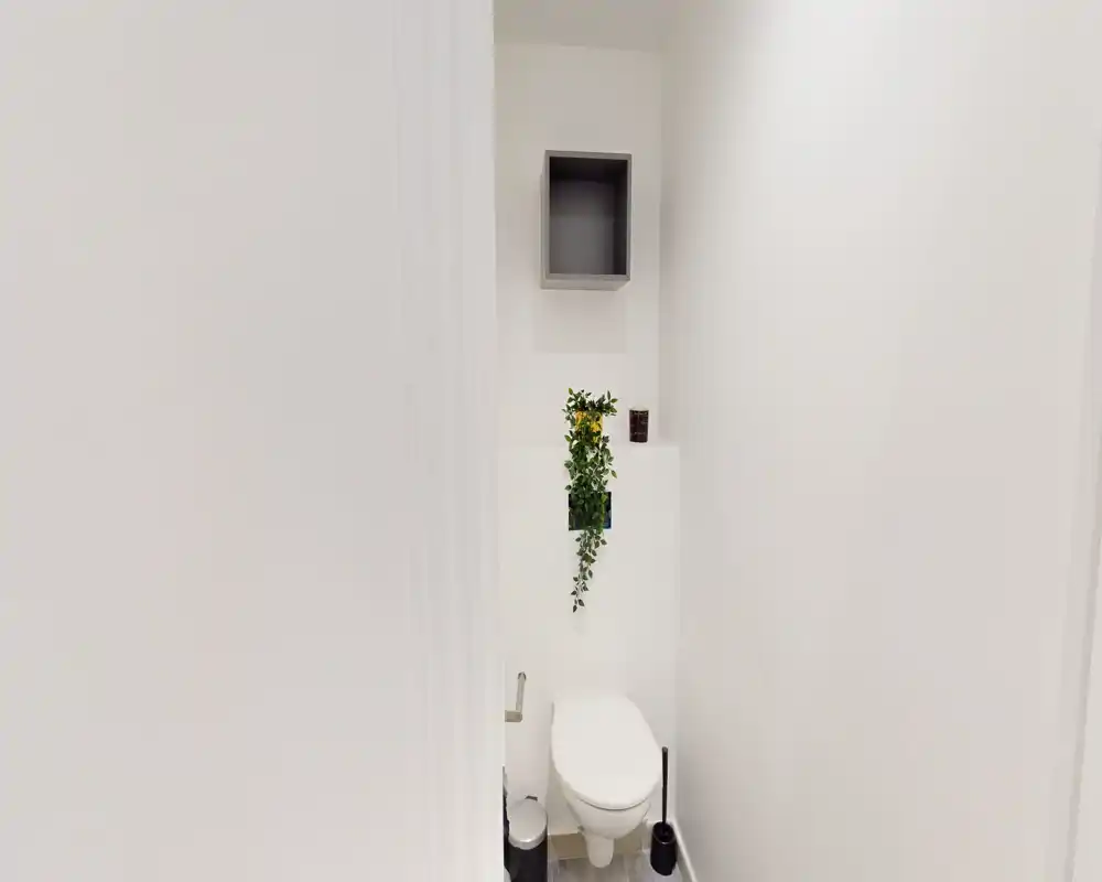 Piccolo bagno con WC compatto, cassettone a muro, mensola decorativa e pianta sospesa; finitura bianca moderna e illuminazione chiara.