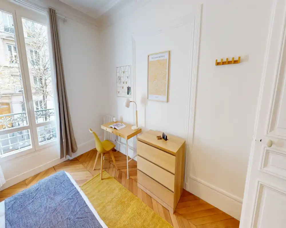 Dormitorio luminoso y bien decorado con suelo de madera, escritorio junto a la ventana, cómoda y cama parcialmente visible con tonos amarillos coordinados.