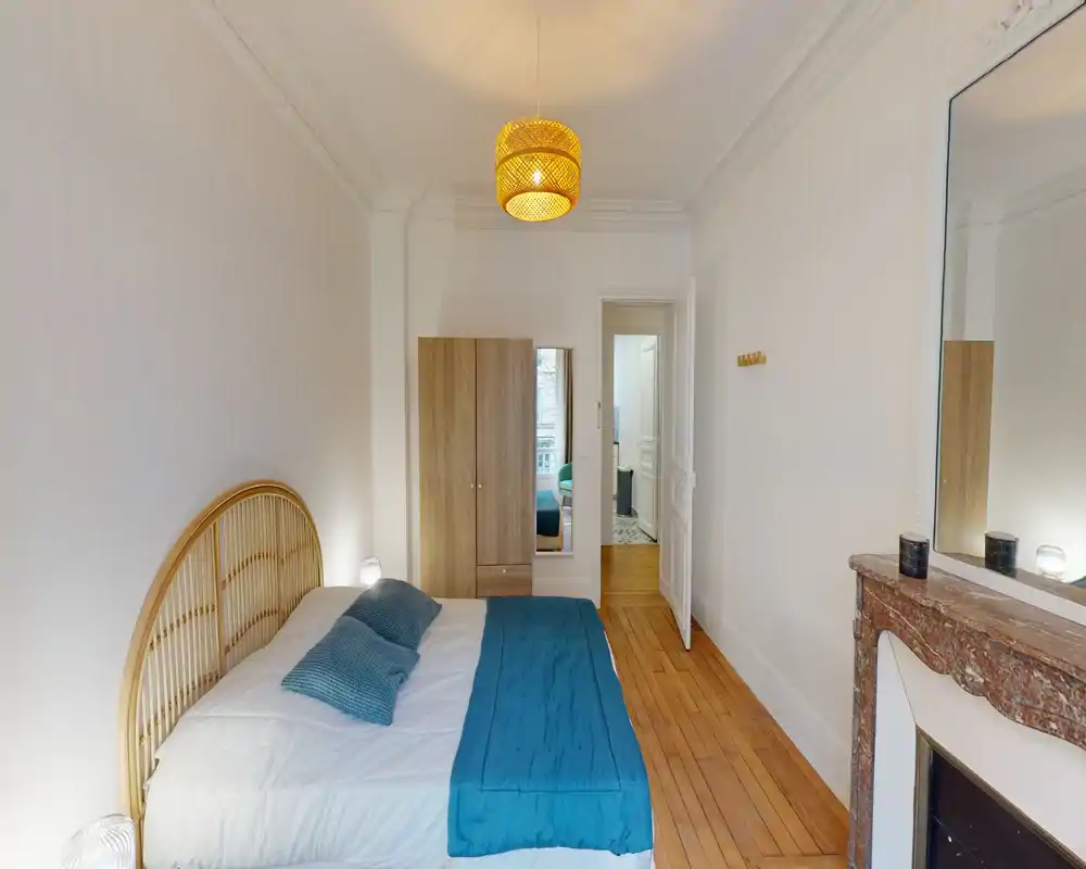Dormitorio luminoso y bien decorado con cabecero de ratán, pie de cama y cojines verde azulado, suelo de madera y chimenea decorativa — perfecto para anuncios de alquiler que destacan un ambiente acogedor.