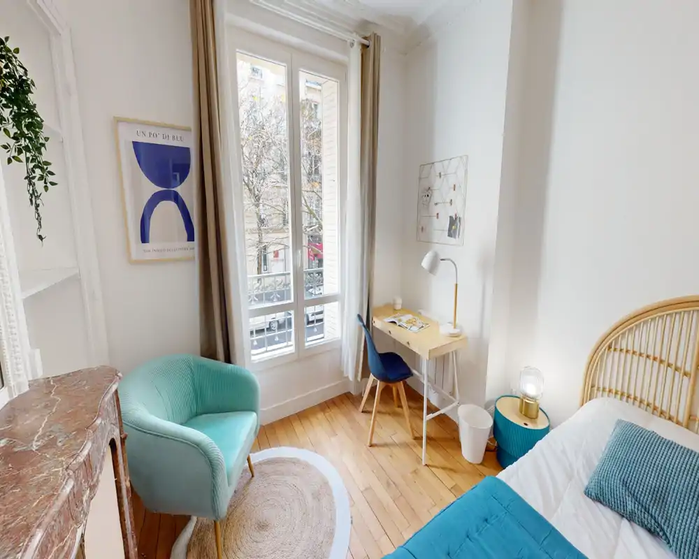 Dormitorio luminoso y acogedor con cabecero de ratán, colcha turquesa y un pequeño escritorio junto a una gran ventana que aporta abundante luz natural.
