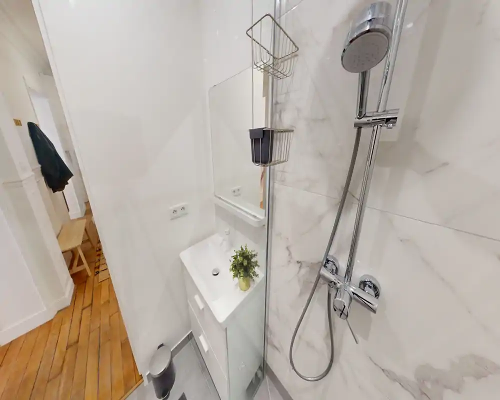 Baño moderno y compacto con ducha, grifería cromada y un pequeño lavabo blanco con espejo. Azulejos efecto mármol y accesorios ordenados ofrecen un aspecto limpio y contemporáneo.