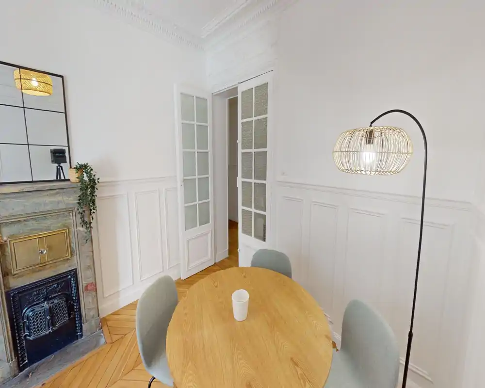 Acogedor rincón comedor luminoso con mesa redonda de madera, sillas tapizadas y lámpara de pie moderna; panelado clásico y chimenea decorativa dan carácter.