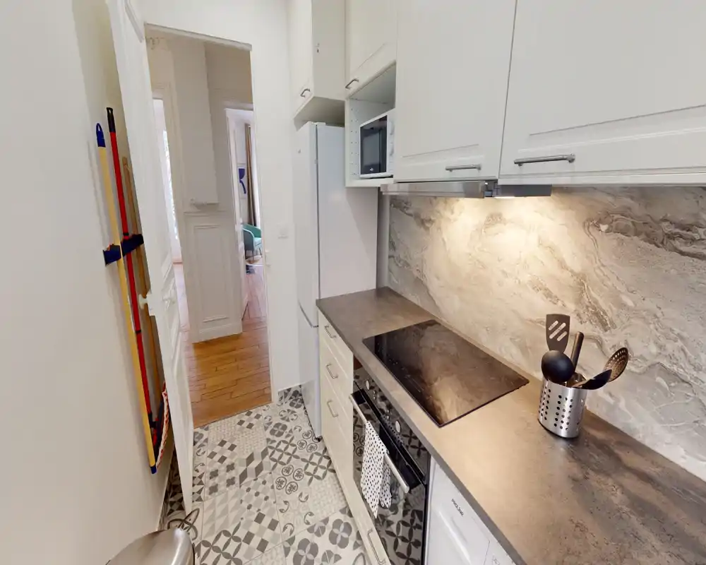 Cocina estrecha y luminosa tipo galería con placa de inducción moderna, salpicadero efecto piedra y suelo de baldosas decorativas. Muebles limpios y encimera práctica, ideal para pisos urbanos.