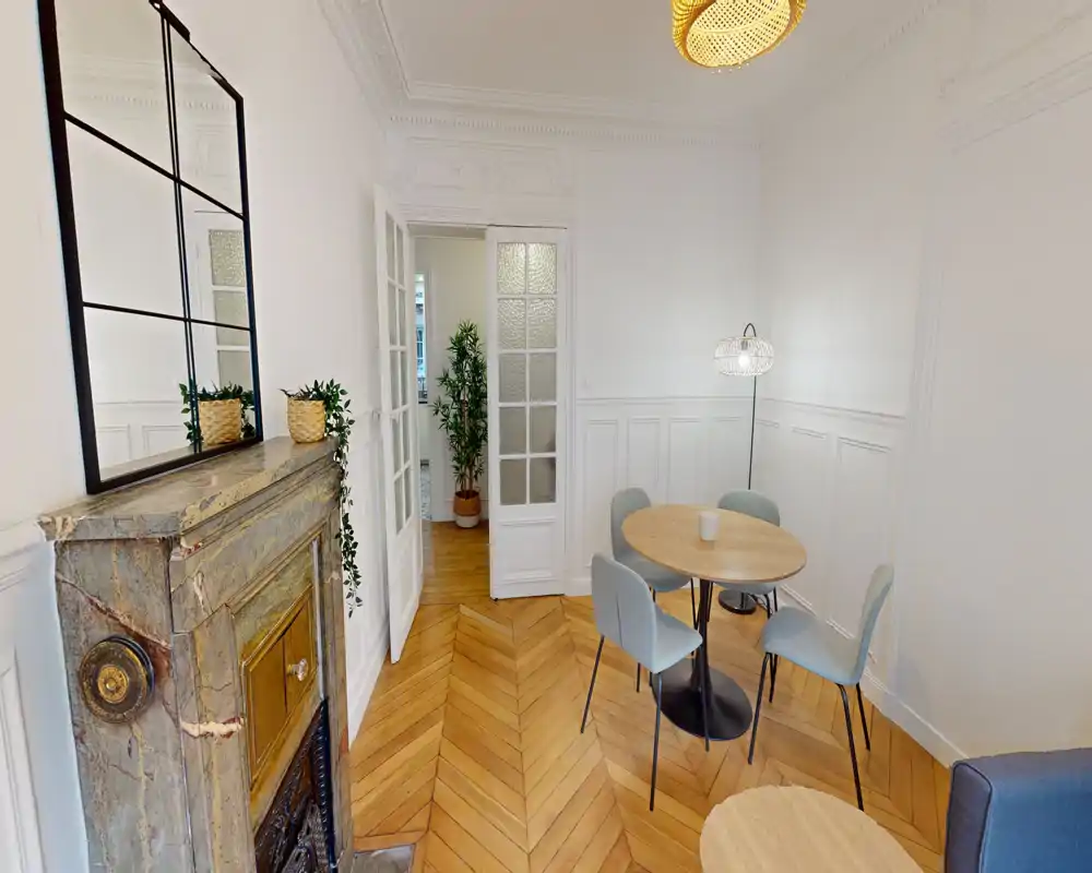 Rincón de comedor luminoso y con estilo con mesa redonda de madera, cuatro sillas modernas y molduras decorativas; el cálido suelo en espiga y una chimenea vintage añaden carácter.
