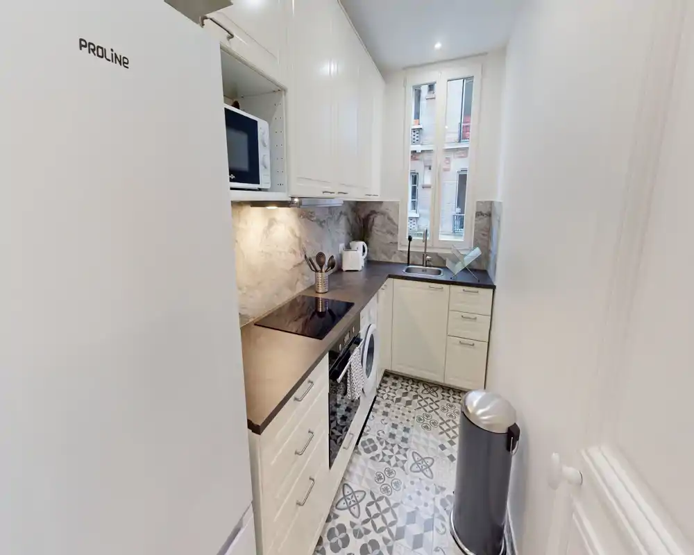 Cocina tipo pasillo luminosa con muebles blancos, suelos con estampado, horno empotrado, placa de inducción y nevera alta — ordenada, moderna y bien iluminada.