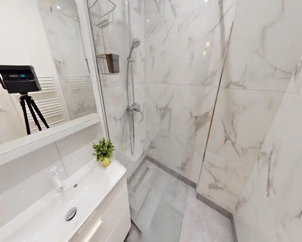 Baño moderno con ducha a ras de suelo, mampara de vidrio, cabezal de ducha de pared y lavabo pequeño con espejo; azulejos claros efecto mármol y suelo gris para un aspecto limpio y contemporáneo.