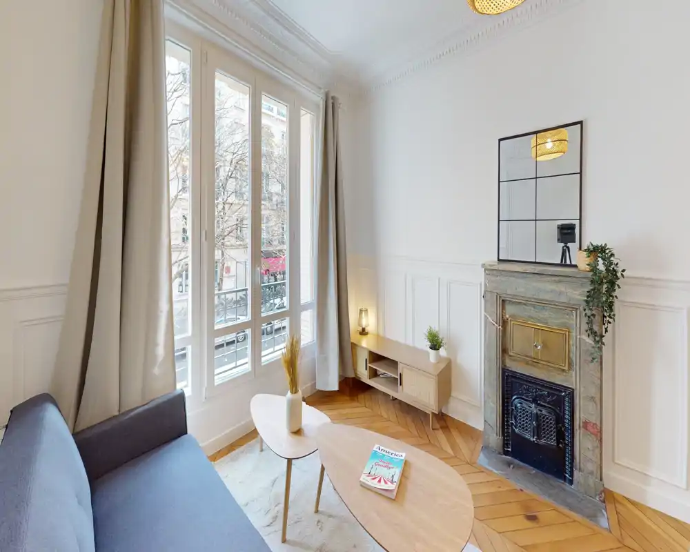 Sala de estar luminosa y con estilo con una gran ventana, sofá acogedor, mesas de madera y una chimenea ornamentada — ideal para alquileres en la ciudad.