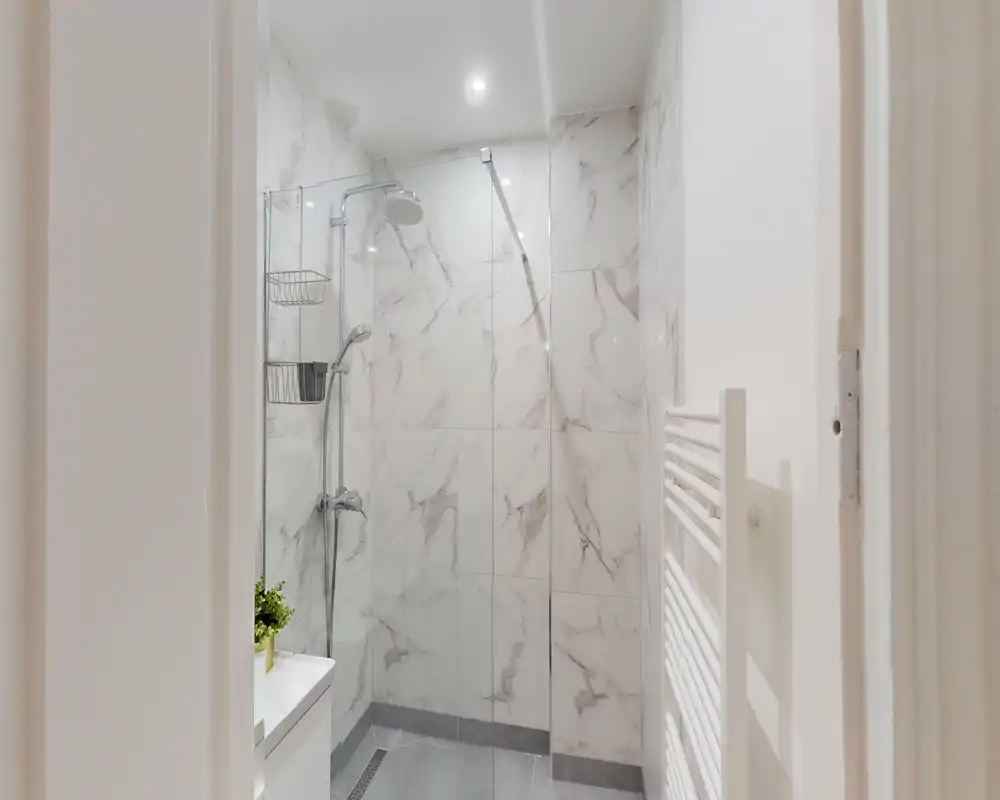 Baño moderno con azulejos tipo mármol, ducha a ras de suelo con panel de cristal, grifería cromada y toallero calefactado; luminoso y limpio.