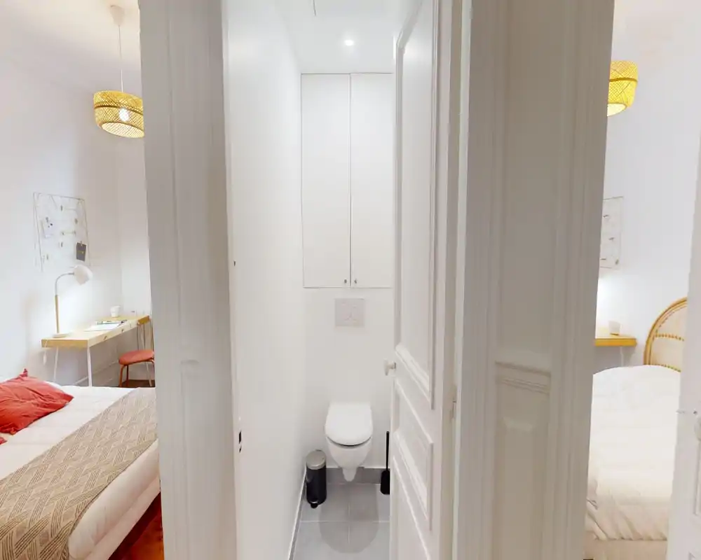 Vista luminosa y simétrica del dormitorio con dos camas individuales a ambos lados de un pequeño baño central: lámparas cálidas, textiles y una zona de trabajo ordenada crean un ambiente acogedor.
