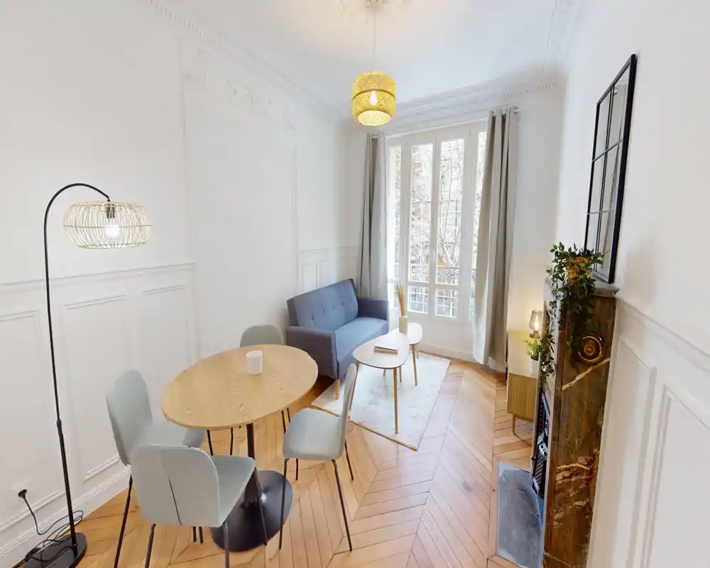Sala luminosa y con estilo con sofá compacto, mesa de centro redonda y comedor pequeño sobre parquet en espiga — ventana amplia y molduras que aportan un toque refinado.