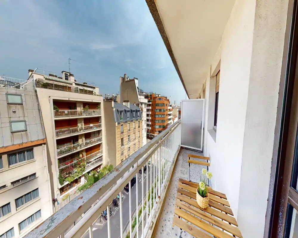 Sunny narrow balcony with a small wooden table and chairs, overlooking a city street and neighboring buildings — ideal for a morning coffee.