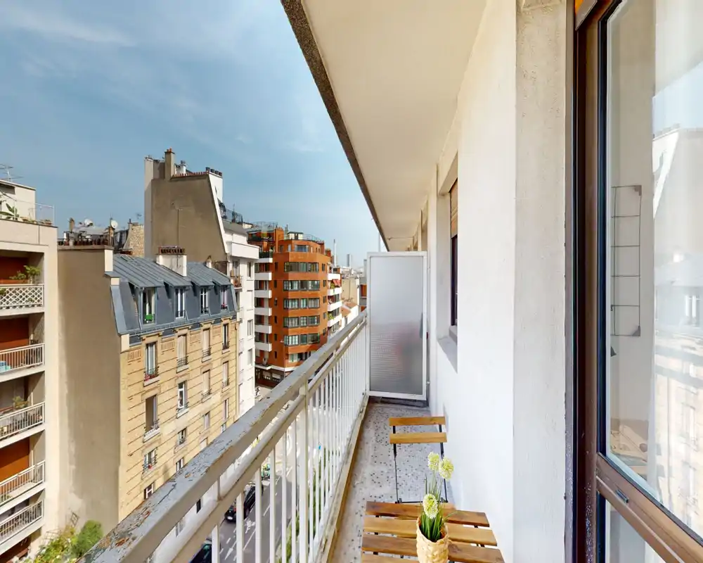 Sunny narrow balcony with a small wooden table and chairs, overlooking a pleasant urban street view. Bright daylight and a neat railing make it suitable for a morning coffee.