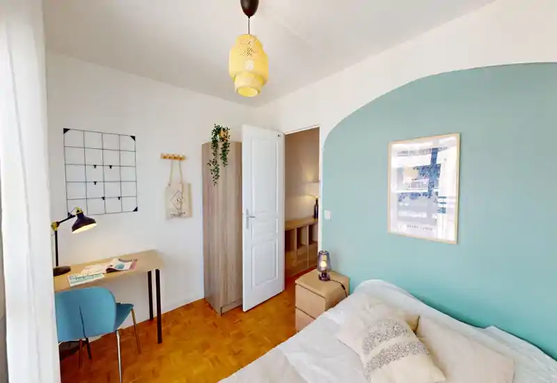Bright, cozy bedroom with a teal accent wall, double bed with cushions, bedside table and lamp, and a small desk by the window — ideal for a comfortable stay or remote work.