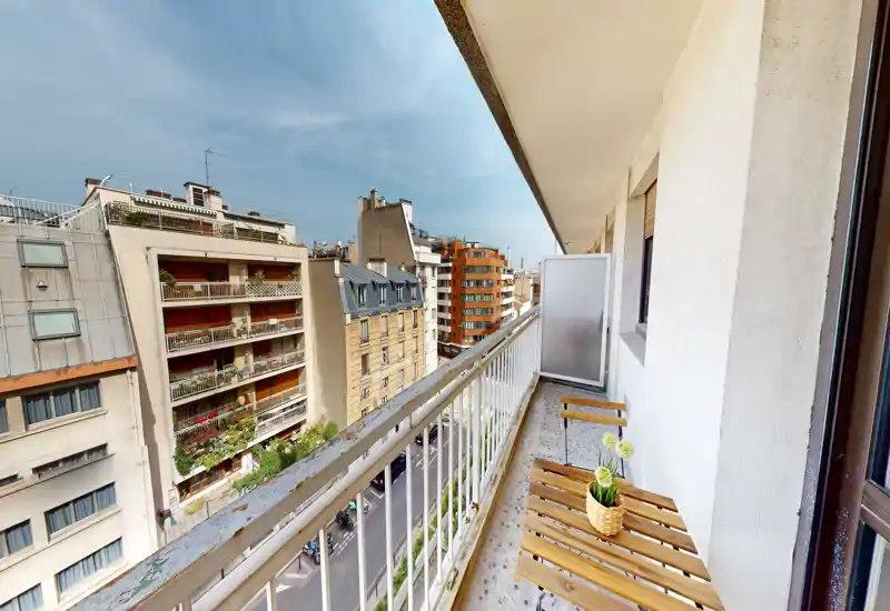 Sunny narrow balcony with a small wooden table and chairs, overlooking a city street and neighboring buildings — ideal for a morning coffee.