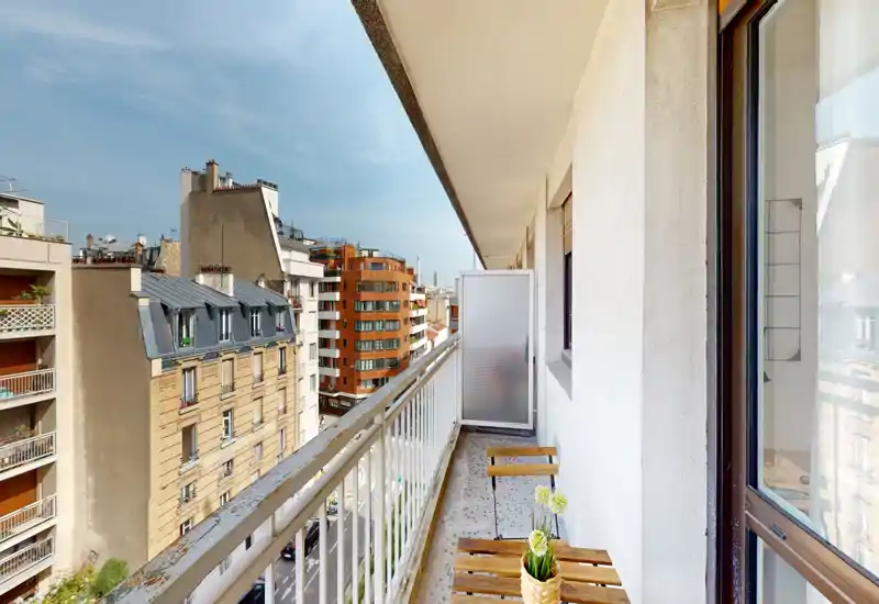 Sunny narrow balcony with a small wooden table and chairs, overlooking a pleasant urban street view. Bright daylight and a neat railing make it suitable for a morning coffee.