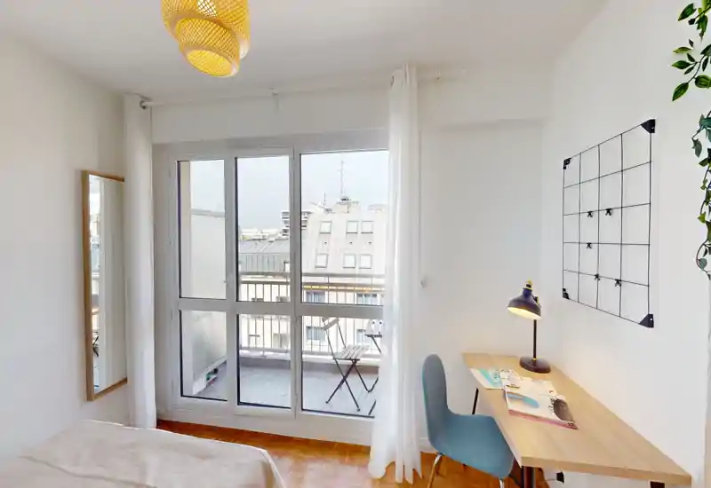 Bright, minimal bedroom with a large balcony window, light wood desk and a comfortable bed partially visible in the foreground — ideal for a city rental with natural light and a small work corner.