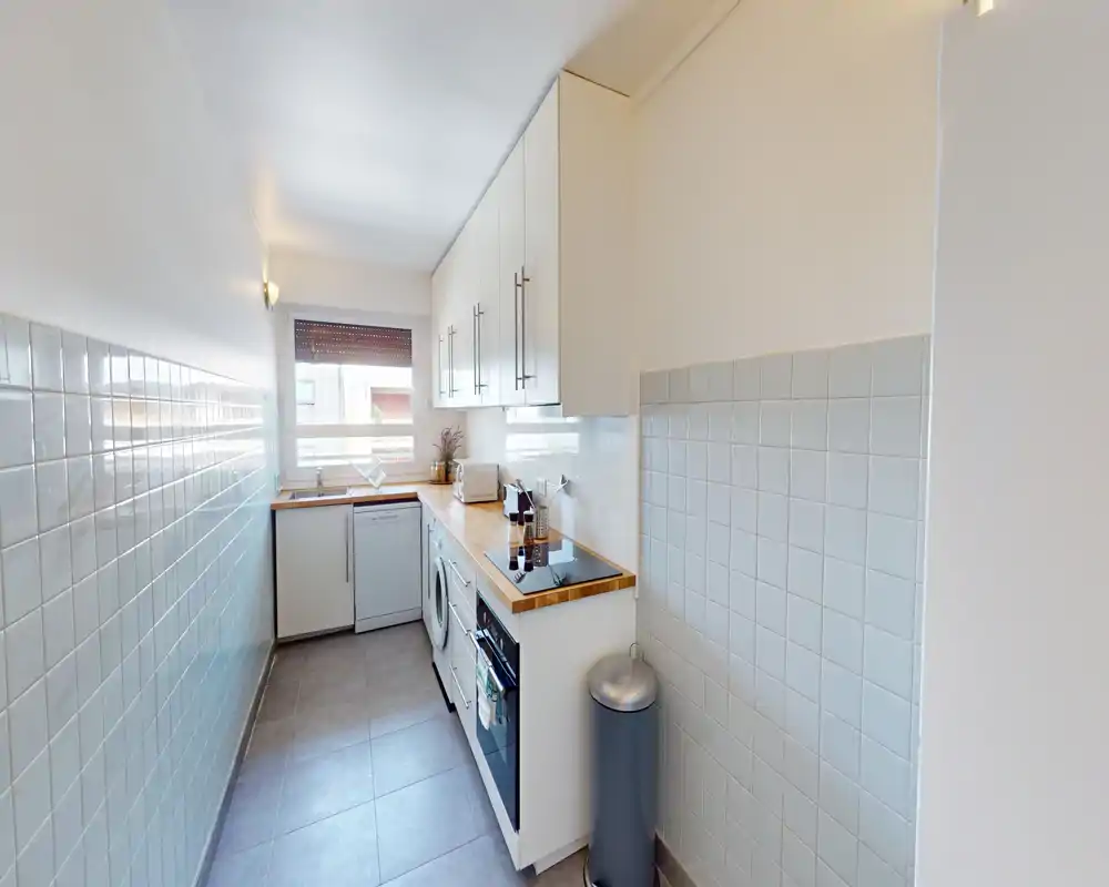 Narrow modern kitchen with white cabinets, wooden countertop, built-in appliances and a bright window providing natural light.