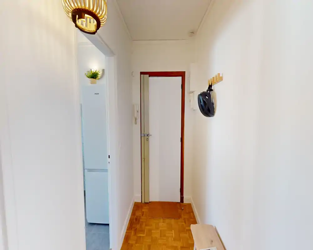 Bright, narrow entrance hallway with wooden parquet floor, wall hooks holding a helmet, and a small bench — clean and welcoming first-impression space.