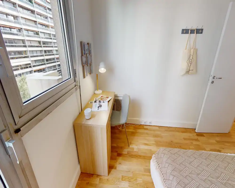 Small, bright bedroom corner with a wooden desk by the window, a bedside mattress with a patterned throw, and a simple chair — neat, minimal and well lit.